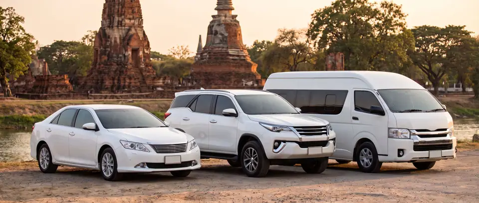 A sedan, an SUV and a van taxi by the river in Ayutthaya, a ruined chedi behind them